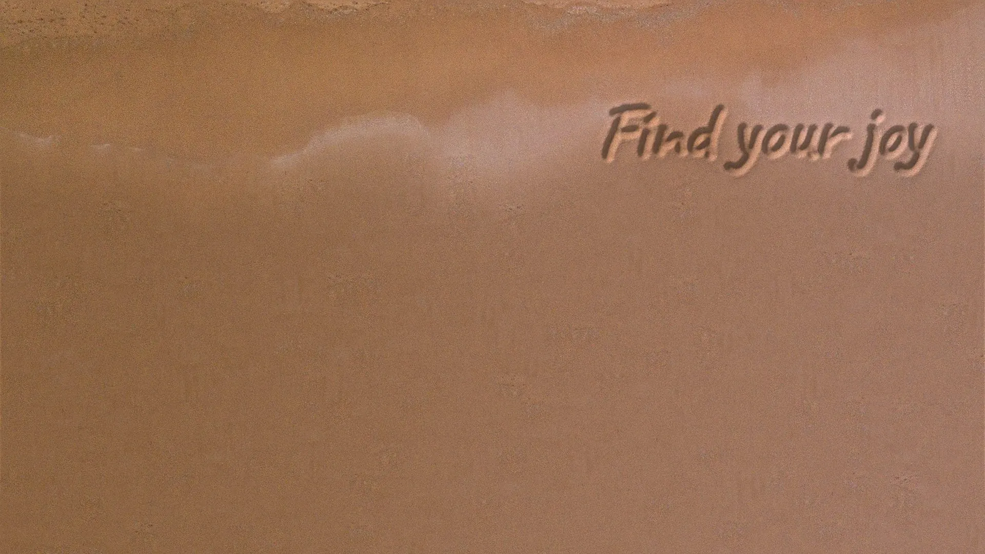 A beach from above: waves washing up the sand, with “Find your joy” written in it